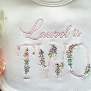 May include: White baby bib with embroidered text "Laurel is TWO" in a delicate pink script. The letters "T" and "W" are adorned with floral embroidery in shades of pink, green, and yellow, creating a charming design.