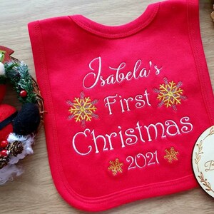 May include: Red baby bib with white embroidered text that reads "Isabela's First Christmas 2021". The bib features yellow snowflake designs. A wooden coaster with the text "BB Threads" is in the lower right corner.