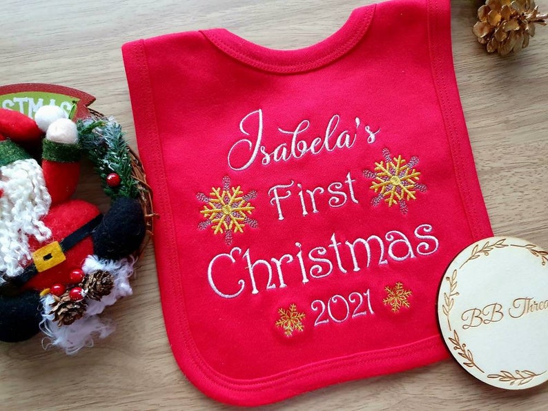 May include: Red baby bib with white embroidered text that reads "Isabela's First Christmas 2021". The bib features yellow snowflake designs. A wooden coaster with the text "BB Threads" is in the lower right corner.