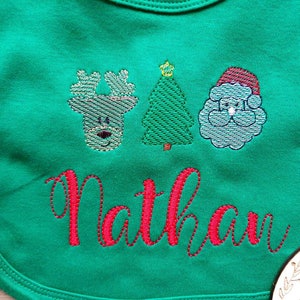 May include: Green baby bib with embroidered Christmas designs: a reindeer, a Christmas tree, and Santa Claus. The name "Nathan" is embroidered in red script below the designs. The bib is made of soft fabric.