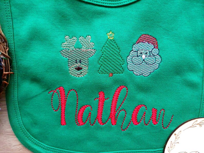May include: Green baby bib with embroidered Christmas designs: a reindeer, a Christmas tree, and Santa Claus. The name "Nathan" is embroidered in red script below the designs. The bib is made of soft fabric.