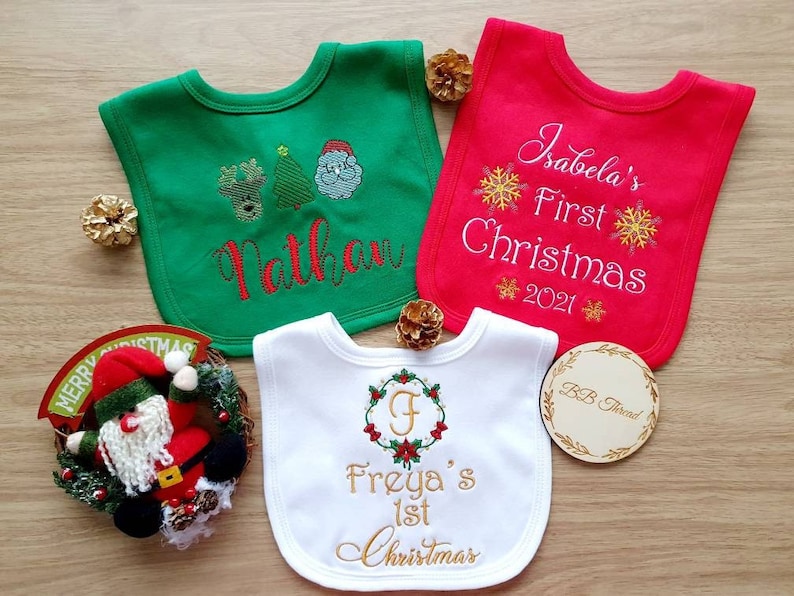 May include: Three baby bibs in green, red, and white, embroidered with Christmas-themed designs and names. The green bib features a reindeer, Christmas tree, and Santa. The red bib says "Isabela's First Christmas 2021". The white bib says "Freya's 1st Christmas".