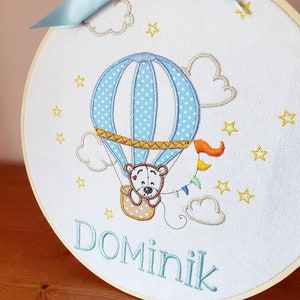 May include: Embroidered hoop art featuring a blue hot air balloon with white polka dots, a teddy bear, and the name "Dominik" in blue thread. The design includes clouds, stars, and a colorful pennant. A light blue ribbon is attached at the top.