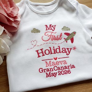 Könnte beinhalten: Weißer Baby-Body mit rosa und roter Stickerei. Der Text lautet "My First Holiday" mit einem Flugzeug, Wolken, dem Namen "Maeva", Ort und Datum. Das Kleidungsstück ist aus weichem Stoff.