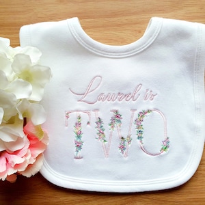 May include: White baby bib with the embroidered text "Laurel is TWO". The letters of "TWO" are decorated with colorful floral embroidery. The bib is a soft, rounded shape, perfect for a toddler.
