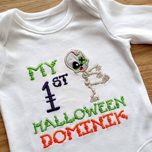 May include: White long-sleeved baby bodysuit with embroidered text and a skeleton design. The text reads "MY 1ST HALLOWEEN DOMINIK" in green, purple, and orange thread. The skeleton has a green eye and is in a dancing pose.