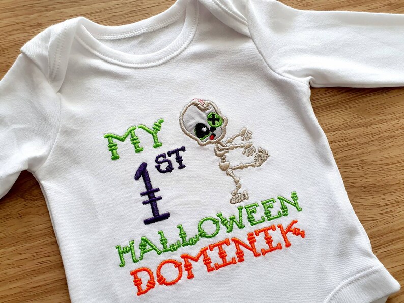 May include: White long-sleeved baby bodysuit with embroidered text and a skeleton design. The text reads "MY 1ST HALLOWEEN DOMINIK" in green, purple, and orange thread. The skeleton has a green eye and is in a dancing pose.
