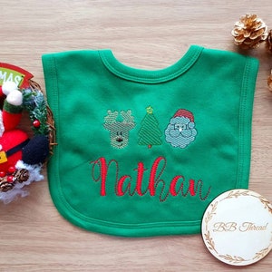 May include: A green baby bib with embroidered Christmas designs, including a reindeer, Christmas tree, and Santa Claus. The name "Nathan" is embroidered in red. A wooden coaster with "BB Thread" is also visible.