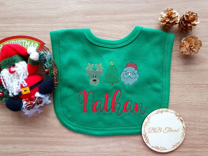 May include: A green baby bib with embroidered Christmas designs, including a reindeer, Christmas tree, and Santa Claus. The name "Nathan" is embroidered in red. A wooden coaster with "BB Thread" is also visible.