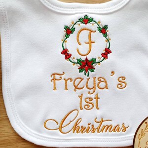 May include: White baby bib with embroidered gold text reading "Freya's 1st Christmas." A Christmas wreath with red and green accents surrounds the letter "F." The bib is made of soft fabric, perfect for a baby's first Christmas.