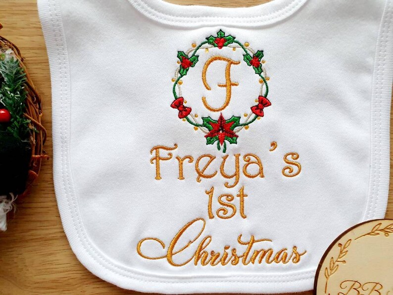 May include: White baby bib with embroidered gold text reading "Freya's 1st Christmas." A Christmas wreath with red and green accents surrounds the letter "F." The bib is made of soft fabric, perfect for a baby's first Christmas.