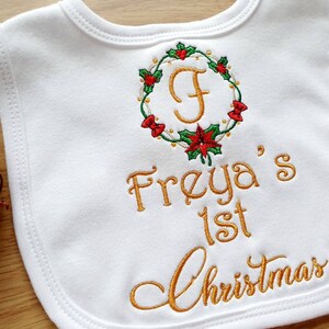 May include: White baby bib with embroidered gold text and a Christmas wreath design. The wreath features red bows, green leaves, and a gold letter "F". The text reads "Freya's 1st Christmas".