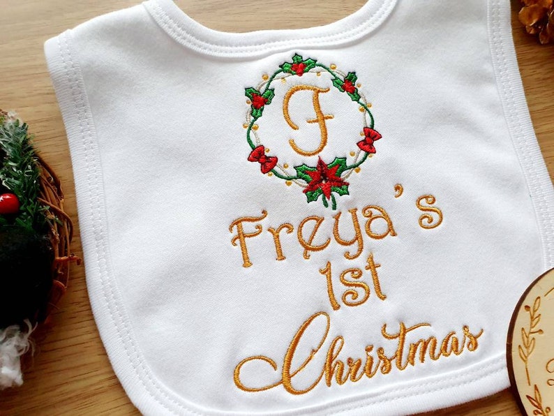 May include: White baby bib with embroidered gold text and a Christmas wreath design. The wreath features red bows, green leaves, and a gold letter "F". The text reads "Freya's 1st Christmas".