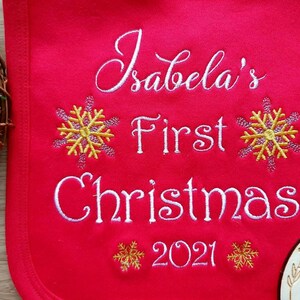 May include: A red bib embroidered with the text "Isabela's First Christmas 2021" in white script. The design includes yellow and pink snowflake accents, perfect for a baby's first holiday season.