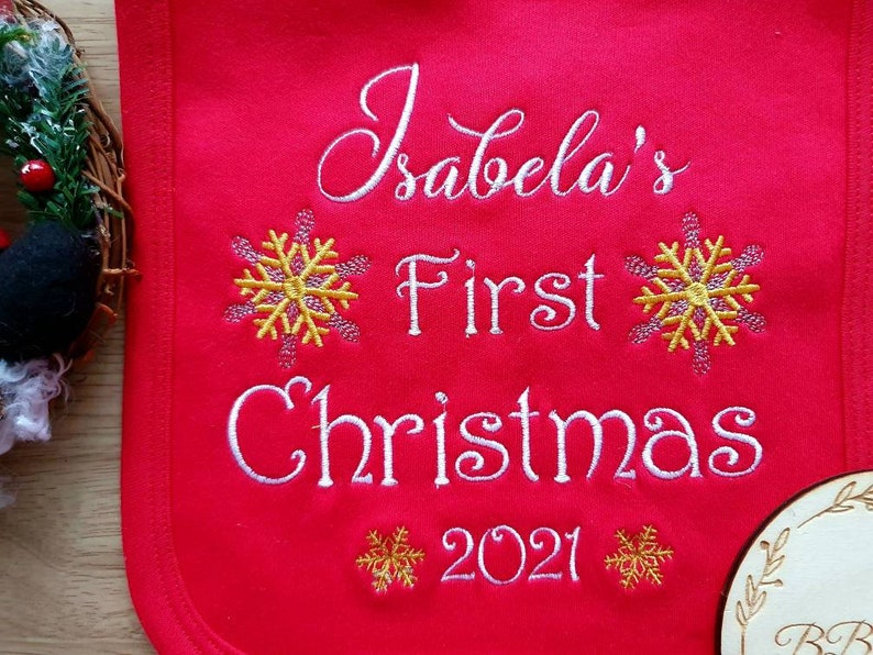 May include: A red bib embroidered with the text "Isabela's First Christmas 2021" in white script. The design includes yellow and pink snowflake accents, perfect for a baby's first holiday season.