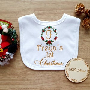 May include: White baby bib with embroidered text "Freya's 1st Christmas" in gold thread. The bib features a wreath design with the letter "F" in the center. A small wooden circle with "BB Thread" is also visible.