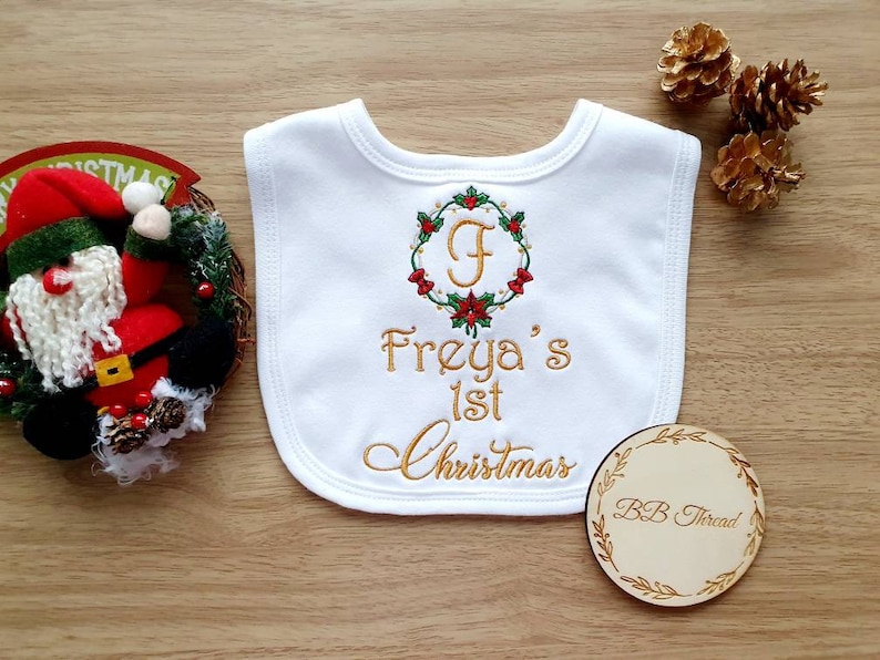 May include: White baby bib with embroidered text "Freya's 1st Christmas" in gold thread. The bib features a wreath design with the letter "F" in the center. A small wooden circle with "BB Thread" is also visible.