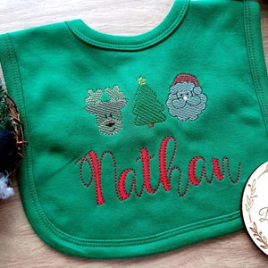 May include: Green baby bib with embroidered Christmas designs, including a reindeer, Christmas tree, and Santa Claus. The name "Nathan" is embroidered in red script. A festive and personalized baby accessory.