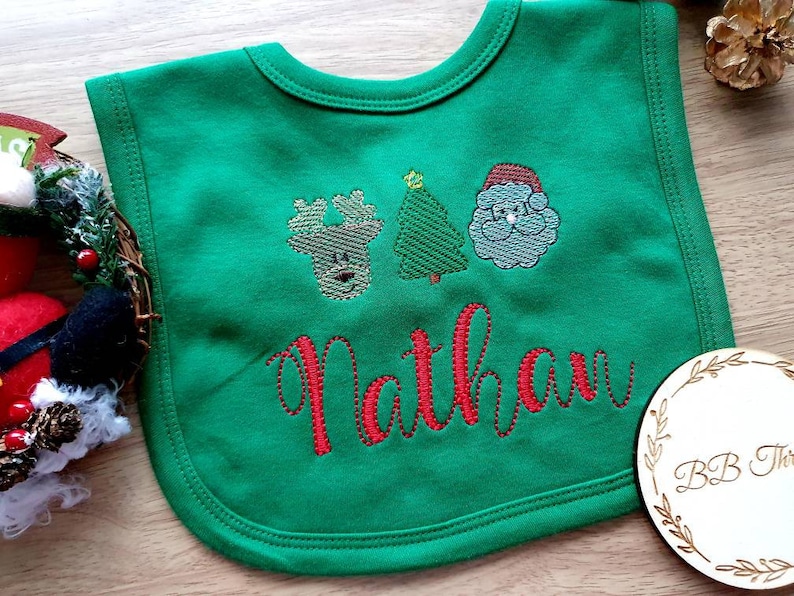 May include: Green baby bib with embroidered Christmas designs, including a reindeer, Christmas tree, and Santa Claus. The name "Nathan" is embroidered in red script. A festive and personalized baby accessory.