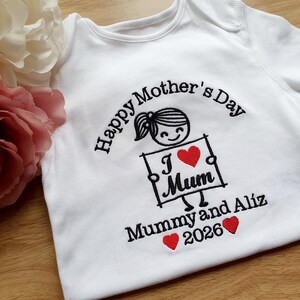 May include: White baby onesie with black embroidered text and a cartoon graphic. The text reads "Happy Mother's Day" and "I Mum" with a heart. Below it says "Mummy and Aliz 2026" with two hearts.