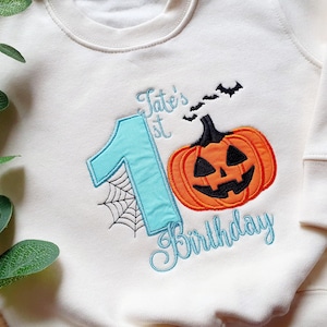 May include: Cream-colored sweatshirt with a Halloween-themed design. Features a large teal number 1 with a spiderweb, an orange jack-o'-lantern, and the text "Tate's 1st Birthday" in teal script. Includes black bats.