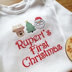 May include: White baby onesie with embroidered Christmas designs. Features a reindeer, Christmas tree, and Santa Claus face above the text "Rupert's First Christmas" in red thread. A wooden round sign is in the background.