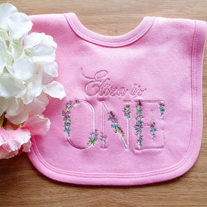 May include: A pink baby bib embroidered with the words "Eliza is ONE." The word "ONE" is decorated with floral embroidery in shades of blue, purple, and green. The bib is made of soft fabric and has a rounded shape.