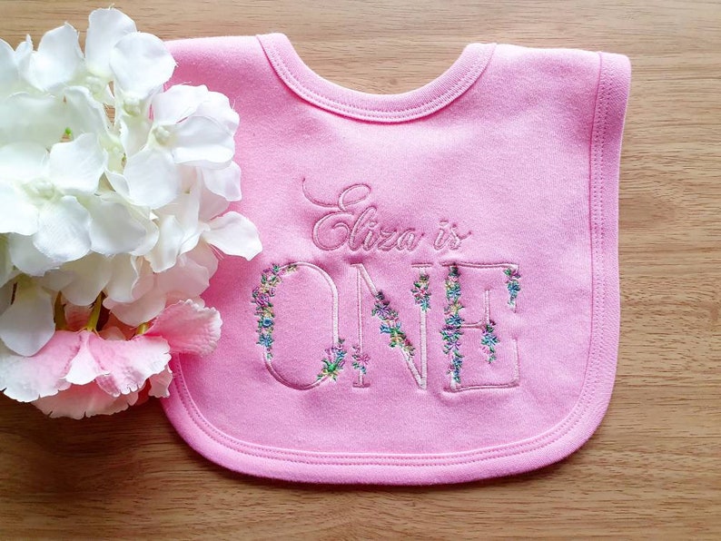 May include: A pink baby bib embroidered with the words "Eliza is ONE." The word "ONE" is decorated with floral embroidery in shades of blue, purple, and green. The bib is made of soft fabric and has a rounded shape.