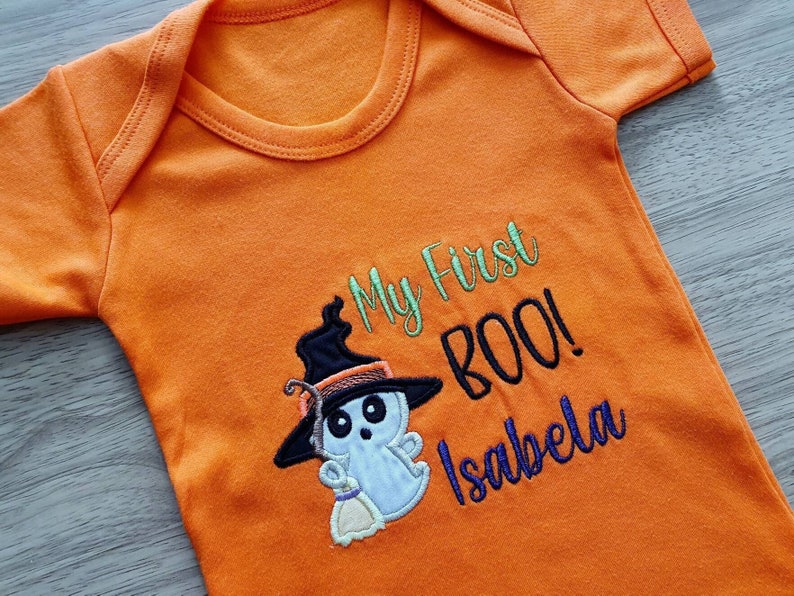 May include: Orange baby bodysuit with the embroidered text "My First BOO!" and the name "Isabela". The design includes a cartoon ghost wearing a witch hat and holding a broom, perfect for Halloween.
