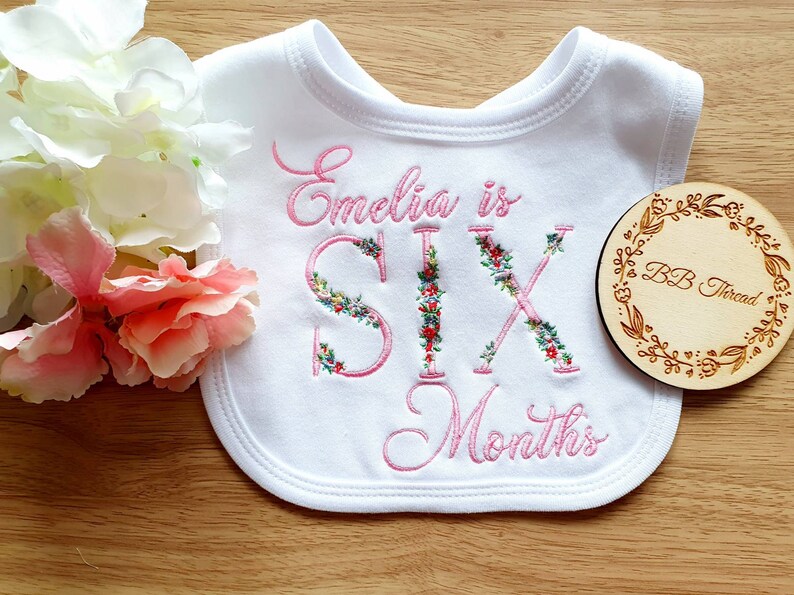 May include: White baby bib embroidered with the words "Emelia is SIX Months" in pink script. The letters of "SIX" are decorated with floral designs. A wooden round with "BB Thread" is next to the bib.