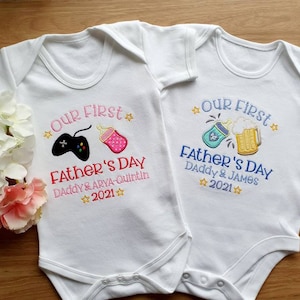 May include: Two white baby onesies with embroidered text and graphics. One features a video game controller and bottle, the other a beer mug and bottle. Both say "Our First Father's Day" and "2021".