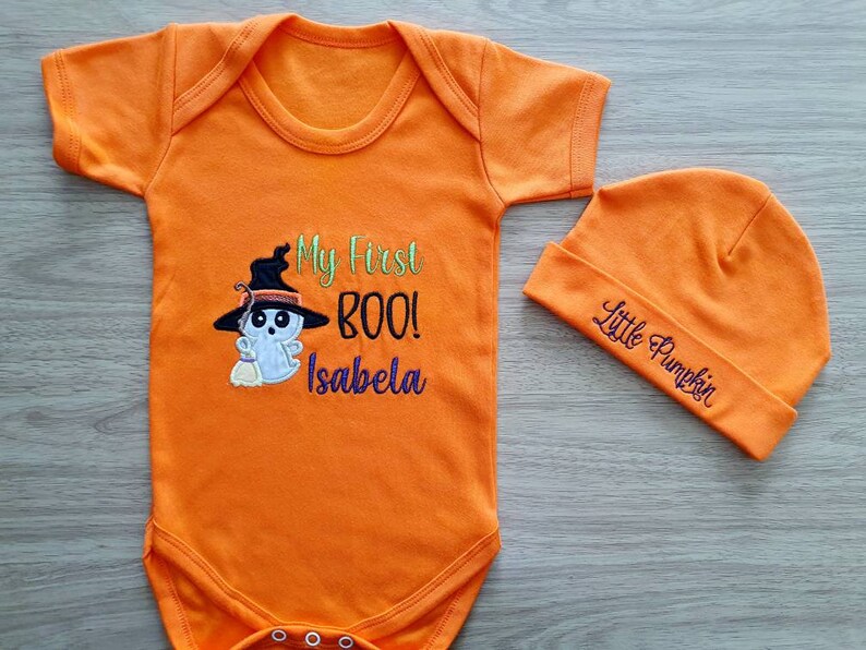 May include: Orange baby bodysuit and beanie set. The bodysuit features an embroidered ghost wearing a witch hat with the text "My First BOO!" and a name. The beanie says "Little Pumpkin". Perfect for Halloween.