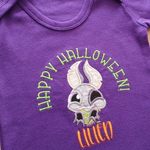 May include: A purple baby onesie with a Halloween-themed embroidered design. The design features a cartoon skull with bunny ears and the words "HAPPY HALLOWEEN!" in green, with the name "LILIEO" in orange.