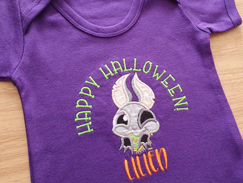 May include: A purple baby onesie with a Halloween-themed embroidered design. The design features a cartoon skull with bunny ears and the words "HAPPY HALLOWEEN!" in green, with the name "LILIEO" in orange.