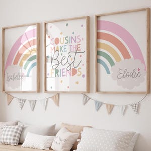 May include: Three framed art prints with rainbow designs and the names "Isabelle" and "Elodie". The central print reads "Cousins Make The Best Friends". The prints are in wooden frames and are decorated with a garland of small flags.