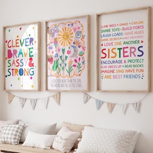 May include: Three framed art prints with positive messages and colourful illustrations. The prints feature phrases like "Clever, Brave, Sassy & Strong," "I love you more than the sun," and "Sisters." A garland of pennants hangs below.