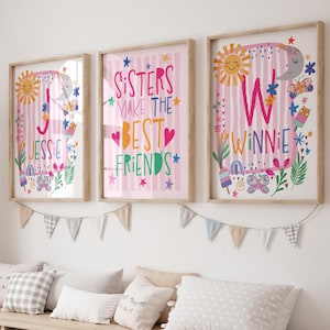 May include: Three framed wall art prints with light wood frames. The left print features the letter J and the name Jessie. The centre print reads Sisters Make The Best Friends. The right print features the letter W and the name Winnie.