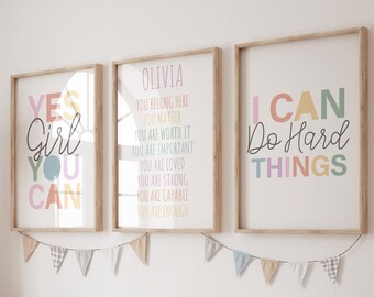 May include: Three framed prints with inspirational quotes hang above a wooden storage bench. The prints feature phrases like "Yes Girl You Can" and "I Can Do Hard Things" in colorful fonts. The bench holds white storage bins and decorative pillows.