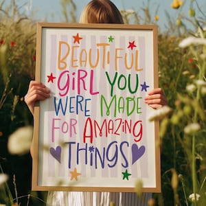 May include: A framed print with the words "BEAUTIFUL GIRL YOU WERE MADE FOR AMAZING THINGS" in colourful lettering. The print has a striped background and is decorated with stars and hearts. The frame is a light brown colour.