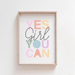 May include: A framed print with the words "YES Girl YOU CAN" in colorful pastel letters. The word "Girl" is in a cursive font, and the other words are in a block font. The frame is a light wood color.