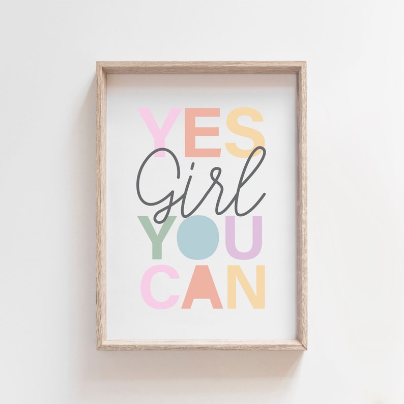 May include: A framed print with the words "YES Girl YOU CAN" in colorful pastel letters. The word "Girl" is in a cursive font, and the other words are in a block font. The frame is a light wood color.