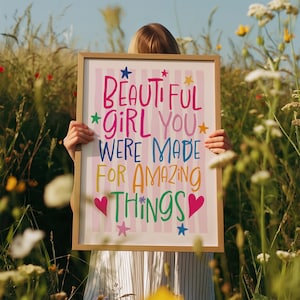 May include: A framed print with the words "BEAUTIFUL GIRL YOU WERE MADE FOR AMAZING THINGS" in colorful lettering. The background has pink and white stripes. The frame is a light brown color. The print is held in a field of tall grass and wildflowers.