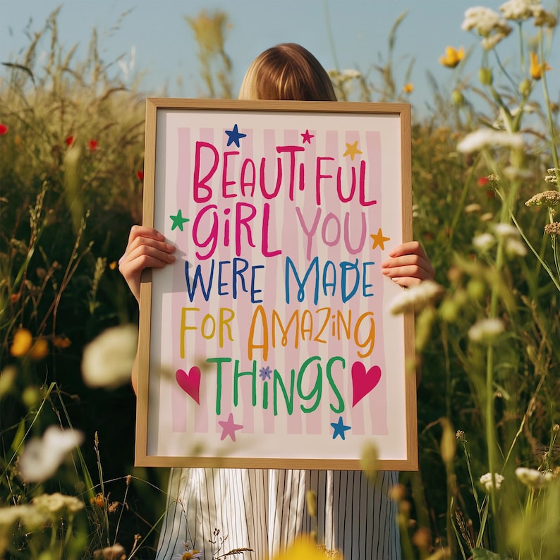 May include: A framed print with the words "BEAUTIFUL GIRL YOU WERE MADE FOR AMAZING THINGS" in colorful lettering. The background has pink and white stripes. The frame is a light brown color. The print is held in a field of tall grass and wildflowers.