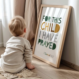 May include: A framed print with the text "FOR THIS CHILD I HAVE PRAYED" in green, gold, and grey lettering. The print is in a light wood frame and is leaning against a wall. A child is sitting on the floor in front of the print.