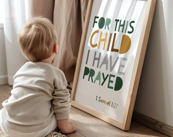 Puede incluir: Un cuadro enmarcado con el texto "FOR THIS CHILD I HAVE PRAYED" en letras verdes, doradas y grises. El cuadro está en un marco de madera clara y está apoyado contra una pared. Un niño está sentado en el suelo frente al cuadro.
