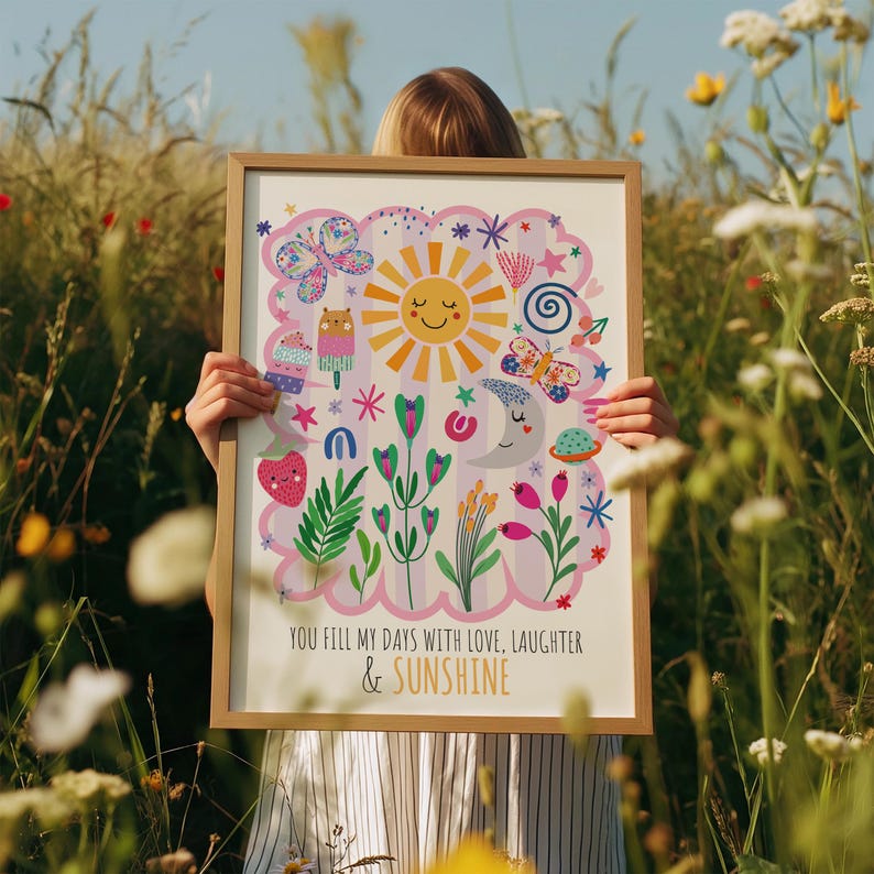 May include: A framed art print featuring a whimsical design with a smiling sun, moon, butterflies, and flowers. The print has a light pink border and includes the text "YOU FILL MY DAYS WITH LOVE, LAUGHTER & SUNSHINE". The frame is a light brown color.