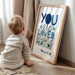 以下が含まれることがあります： 青と緑で「YOU are LOVED beyond MEASURE」という言葉が書かれた額入りのプリント。 プリントは明るい木製のフレームに入っており、壁に立てかけられています。 子供が床に座ってプリントを見ています。