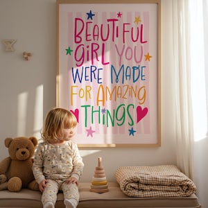 May include: A framed print with the words "BEAUTIFUL GIRL YOU WERE MADE FOR AMAZING THINGS" in colorful lettering hangs above a bench. A young child in a floral onesie sits on the bench next to a teddy bear, a wooden stacking toy, and a folded blanket.
