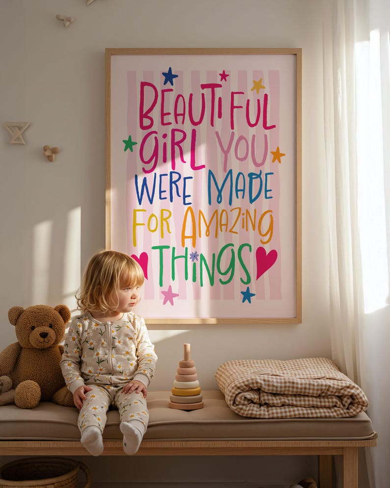 May include: A framed print with the words "BEAUTIFUL GIRL YOU WERE MADE FOR AMAZING THINGS" in colorful lettering hangs above a bench. A young child in a floral onesie sits on the bench next to a teddy bear, a wooden stacking toy, and a folded blanket.