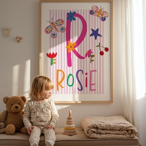May include: A framed print with the name "Rosie" in colorful letters, decorated with butterflies, flowers, and stars. A young child in a floral onesie sits on a bench with a teddy bear, a wooden stacking toy, and a folded blanket.